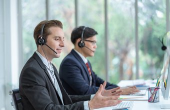 Zwei Männer mit Headsets sitzen nebeneinander am Schreibtisch und arbeiten am Computer.